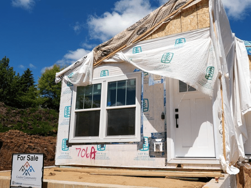 A small in-construction sustainable home is shown at a medium shot on a bright spring day in Hillsboro, Wisconsin. Interior plywood is visible, as is the housewrap around the windows and front door. A small "For Sale" sign is planted in the leftmost foreground.