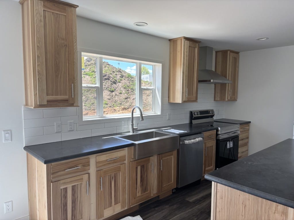A newly furnished and empty kitchen of one of the new factory-built homes in Hillsboro, Wisconsin. The cabinets and drawers are made of wood, while the sink, dishwasher, stove, and overhead fan are stainless steel. Bright natural light shines in through a large window above the sink.