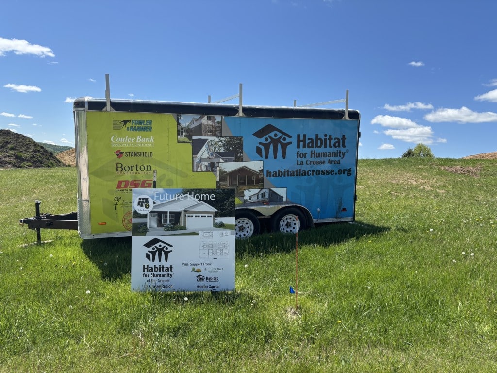 A trailer displaying information for Habitat for Humanity and future homes in the La Crosse, Wisconsin, area sits in a grassy field on a bright spring day.