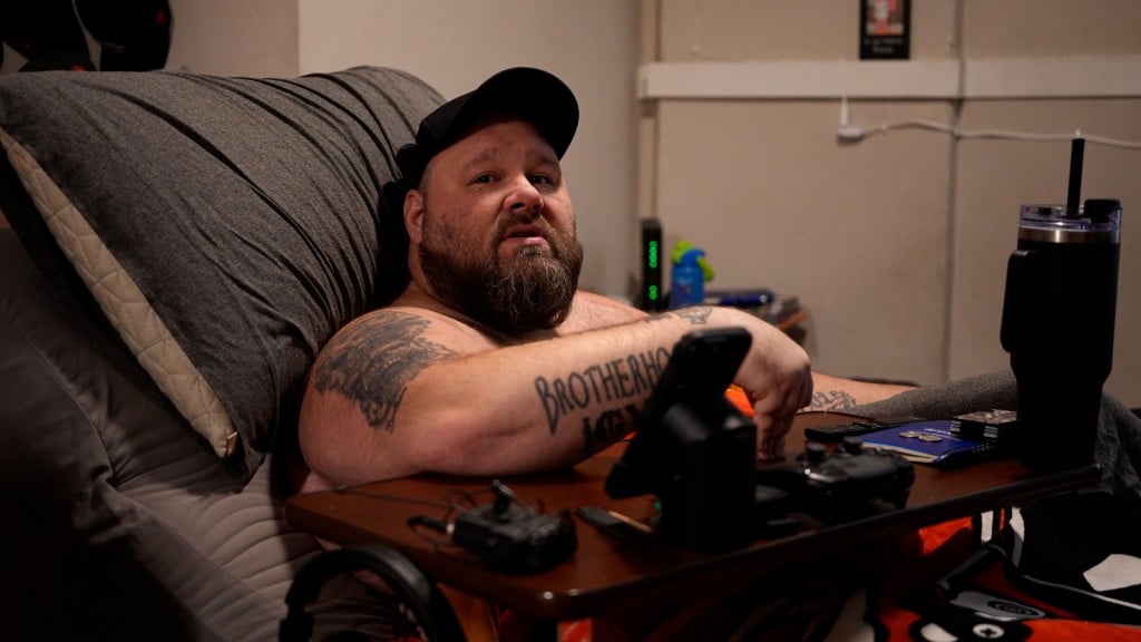 A middle-aged man with a beard sits in a cushioned chair and looks towards the photographer. He wears a dark baseball cap. A wooden lap tray is positioned in front of him. His arms have several tattoos, including one on his right arm that reads "BROTHERHOOD."
