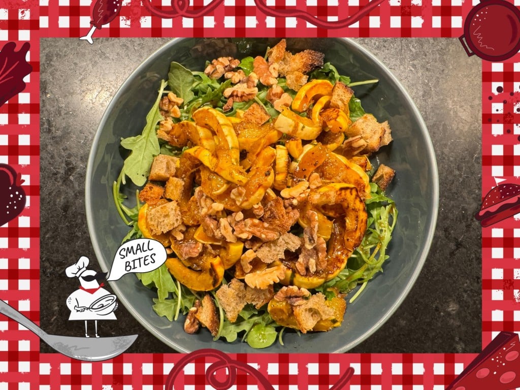 Overhead view of a mixed salad in a grey-green bowl on a dark marble countertop. The bowl contains slices of roasted squash, walnuts, arugula, and croutons. Bordering this photo is a red and white checkered tablecloth illustration. In the lower left corner of this illustrated frame is a small chef with an oversized mustache standing on a spoon. The chef's speech bubble reads "Small Bites."