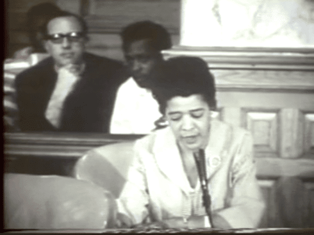 A low-resolution still from the documentary "Decade Of Discontent" shows a Black woman in a suit sitting at a desk in an assembly room, speaking into a microphone. Two men, one white and one Black, sit behind her on the floor and look on.