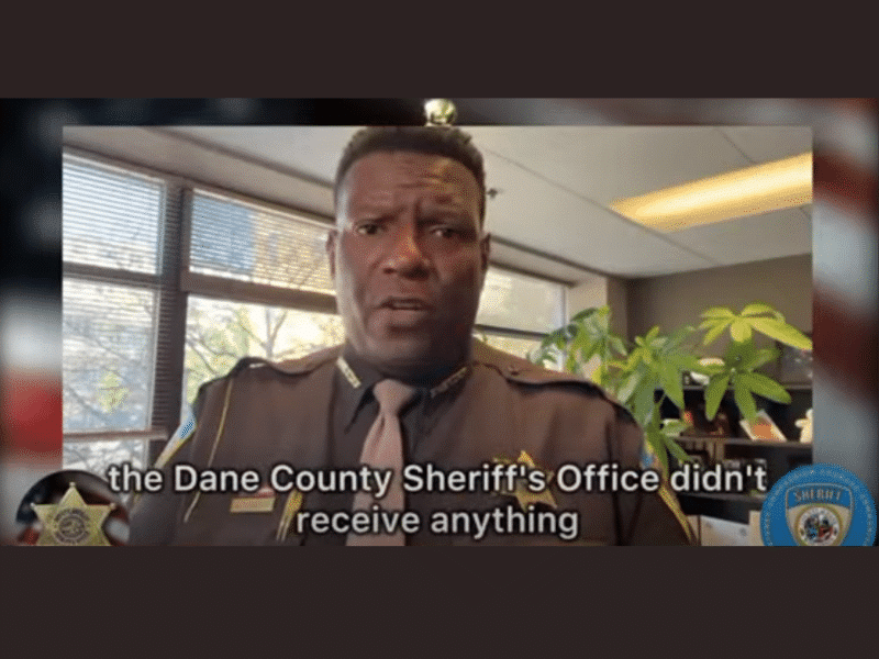 A cropped screenshot from a standard-definition online video shows a Black sheriff in uniform speaking directly into the camera. He stands in his office with bright natural light streaming in through the blinds behind him. A blurry American flag borders the video image, along with two sheriff badge seals along the bottom corners. Subtitles appear in between: "the Dane County Sheriff's Office didn't receive anything."