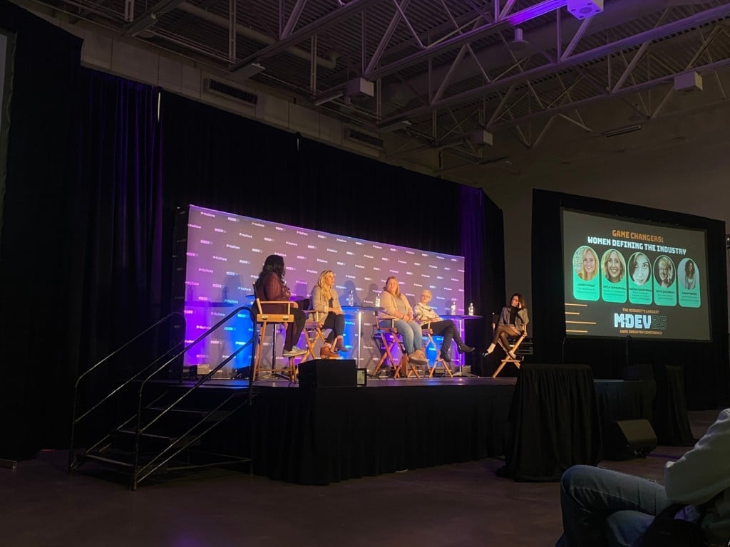 A photograph at a slightly angled long shot of an all-women panel at the MDEV games conference at the Alliant Energy Center in Madison. The hall is rather dimly lit except for the spotlit stage where five women are sitting on elevated chairs. A large screen to the far right of the stage displays the names and photos of the people involved as well as the title "Game Changers: Women Defining The Industry."