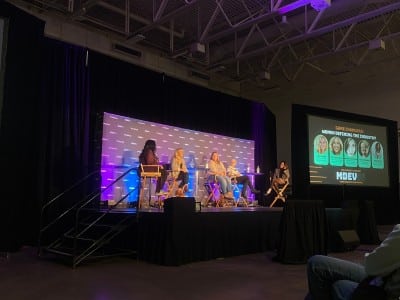A photograph at a slightly angled long shot of an all-women panel at the MDEV games conference at the Alliant Energy Center in Madison. The hall is rather dimly lit except for the spotlit stage where five women are sitting on elevated chairs. A large screen to the far right of the stage displays the names and photos of the people involved as well as the title "Game Changers: Women Defining The Industry."