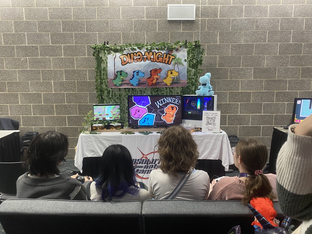 Photo at a medium shot behind four people who are sitting on a couch playtesting a video game with controllers. The table, a few feet in front of them, has three different-sized monitors and a large, colorful promotional sign that hangs above them on the wall. It reads "Dino-Might" and displays illustrations of four baby dinosaurs in green, blue, red, and yellow colors.