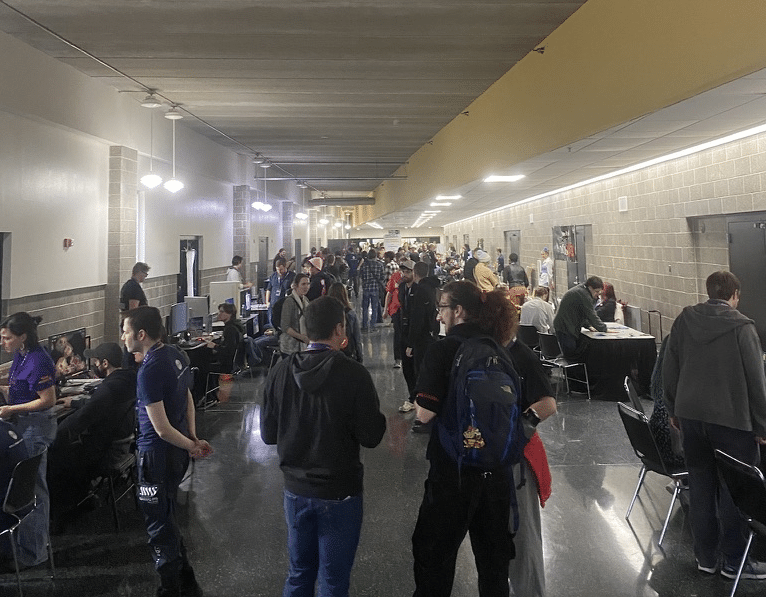 Candid photo of a large, bustling hallway at the Alliant Energy Center in Madison. Dozens of people stand around talking in the middle of the hall or sitting and playtesting video games at tables along the walls.