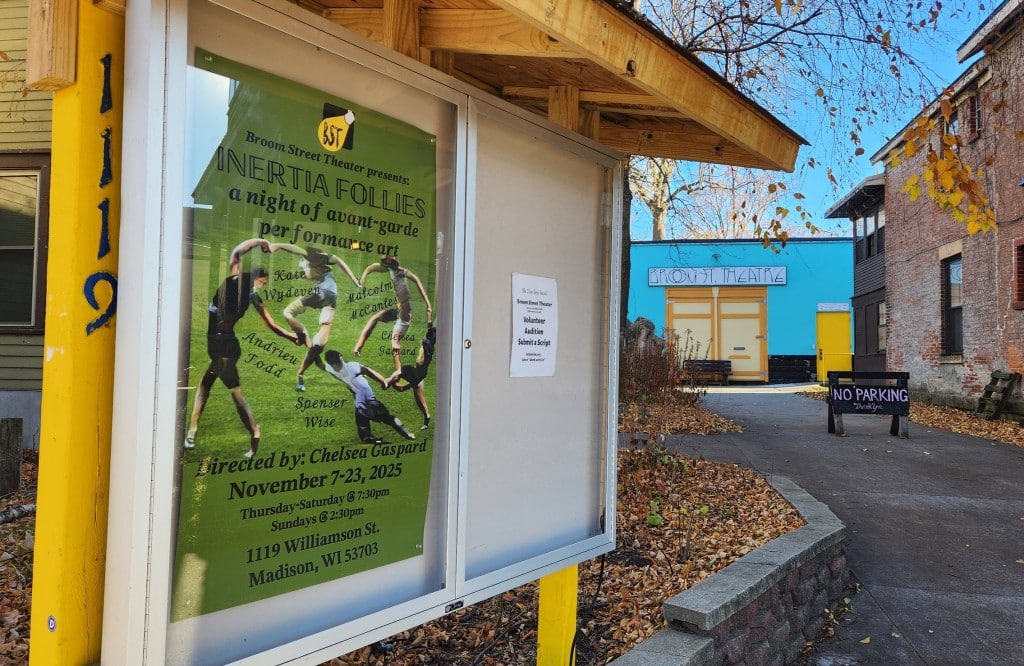 Autumn season photograph of the cased bulletin board in front of Broom Street Theater in Madison at 1119 Williamson Street. The case displays the predominantly forest green-colored show poster with information about "Inertia Follies." The yellow-door entrance to the theater can be seen in the background at the end of a concrete walking path and wooden sign that reads "No Parking."