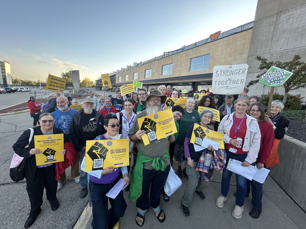 Photo of a few dozen Group Health Cooperative employees and union supporters of various ages gathered outside the GHC building. They pose with signs for the camera. Many of the signs feature a yellow background with the solidarity fist and inspiring language about standing with GHC workers and unions. A few other signs display "Stronger Together" and "Unions Benefit Every One."