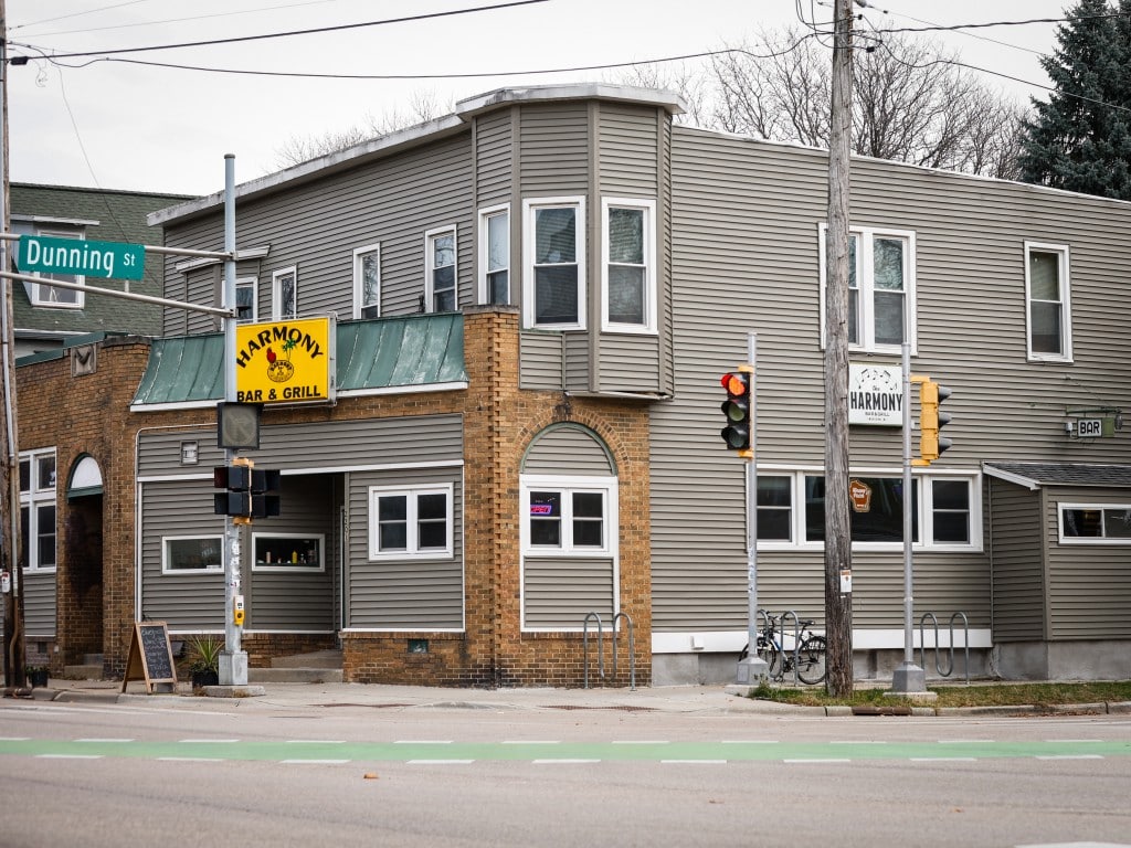 The exterior of Harmony Bar & Grill on the corner of Dunning Street and Atwood Avenue in Madison, WI. The building has gray siding, a yellow marquee sign with a central yellow sign. Muted green accents and a classic brick facade surrounds the gray paneling. A street sign is visible, and the road around the venue is empty.