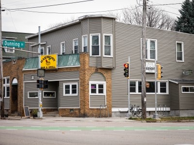 The exterior of Harmony Bar & Grill on the corner of Dunning Street and Atwood Avenue in Madison, WI. The building has gray siding, a yellow marquee sign with a central yellow sign. Muted green accents and a classic brick facade surrounds the gray paneling. A street sign is visible, and the road around the venue is empty.