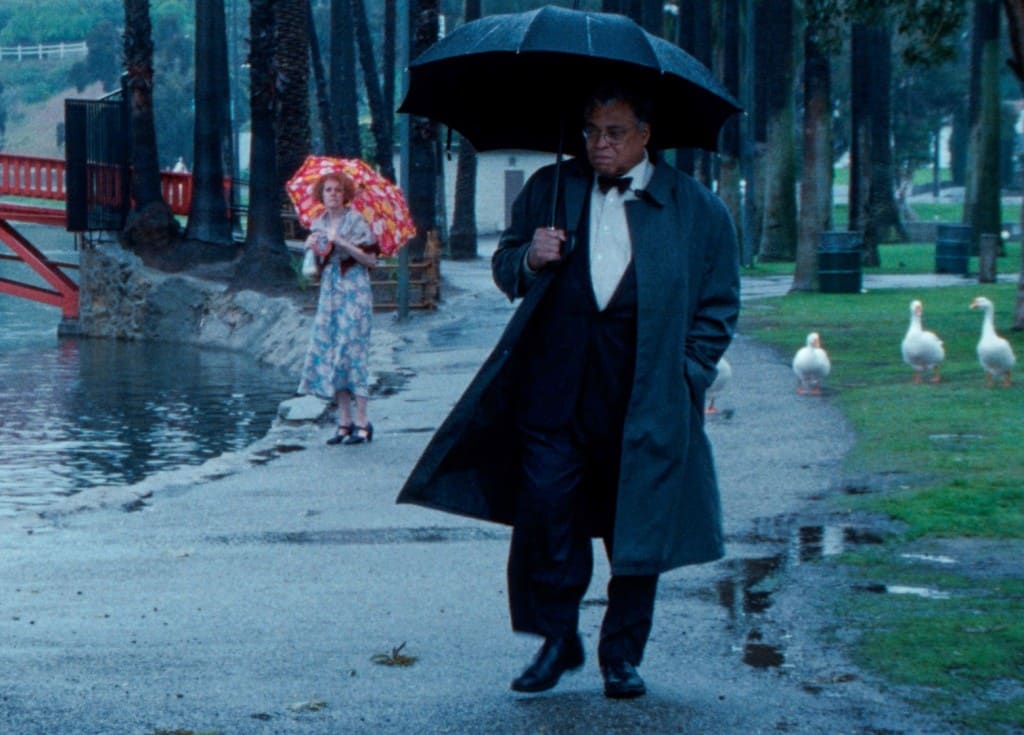 Still image from the film "The Annihilation Of Fish" shows a rainy park scene in Los Angeles with geese along the grass and paved trail. A man in a tuxedo and black trenchcoat carries a black umbrella in the foreground. His facial expression appears pensive and perhaps a bit upset. In the middleground, a woman in a blue floral dress and red and orange floral umbrella stands still and looks at him with a sense of sympathy and concern.