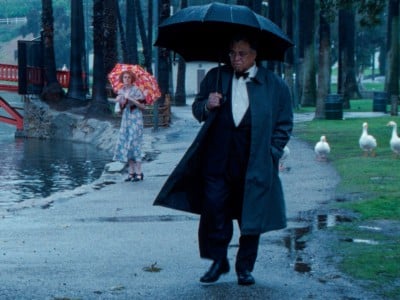Still image from the film "The Annihilation Of Fish" shows a rainy park scene in Los Angeles with geese along the grass and paved trail. A man in a tuxedo and black trenchcoat carries a black umbrella in the foreground. His facial expression appears pensive and perhaps a bit upset. In the middleground, a woman in a blue floral dress and red and orange floral umbrella stands still and looks at him with a sense of sympathy and concern.