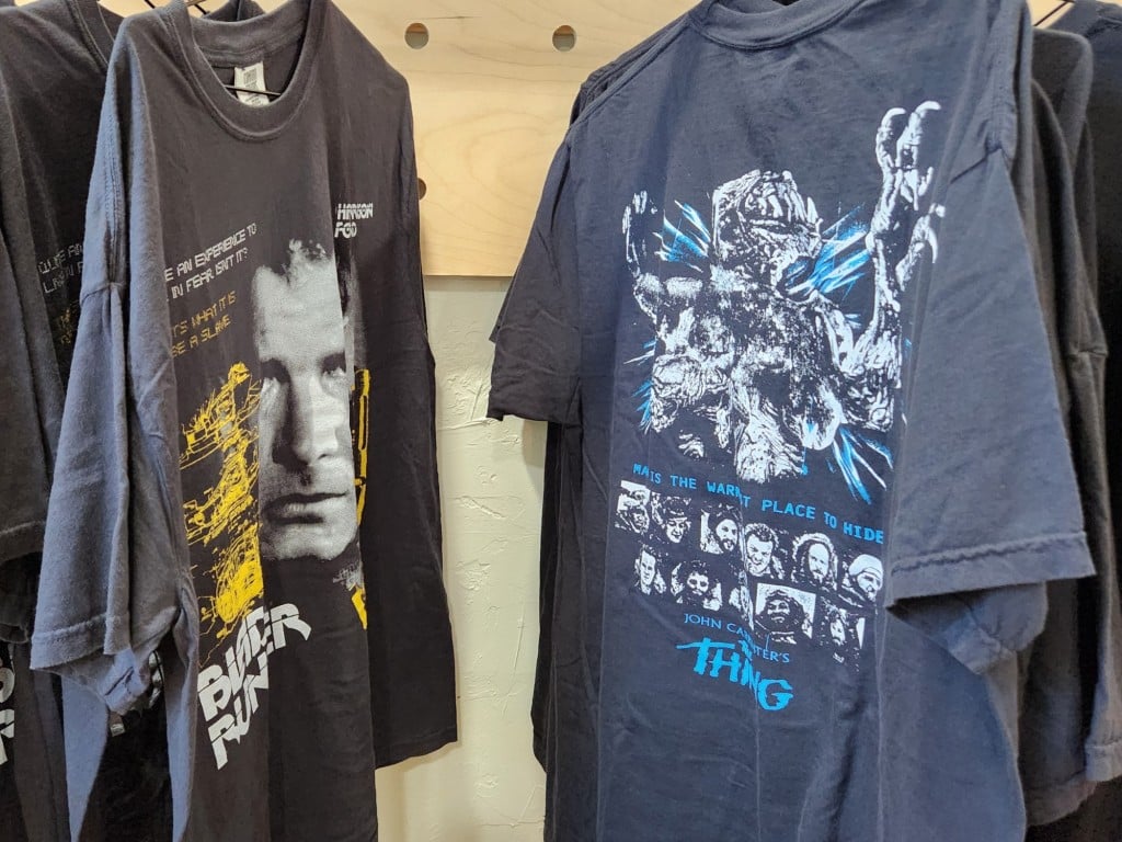 A photograph shows a medium close-up of different sizes of two t-shirt designs hanging on a clothing rack. The leftmost one is "Blade Runner" and the rightmost one is "The Thing." The "Blade Runner" tee prominently features Deckard's face (Harrison Ford) as well as text from the film in yellow and white, while "The Thing" tee includes small portraits of the cast arranged in two long rows with blue text and the iconic alien monster design rendered in black and white.
