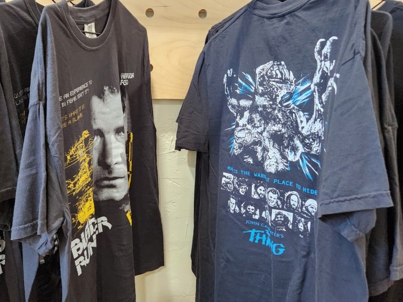 A photograph shows a medium close-up of different sizes of two t-shirt designs hanging on a clothing rack. The leftmost one is "Blade Runner" and the rightmost one is "The Thing." The "Blade Runner" tee prominently features Deckard's face (Harrison Ford) as well as text from the film in yellow and white, while "The Thing" tee includes small portraits of the cast arranged in two long rows with blue text and the iconic alien monster design rendered in black and white.