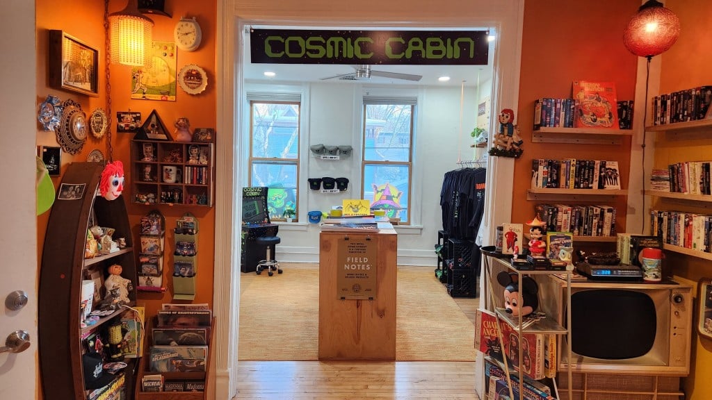 A photo shows various collectible paraphernalia—toys, records, VHS tapes, board games, etc.—along the walls in a mood-lit foyer. In the center is a wide entrance that opens into a brighter sunlit room that extends into the background. The back room displays racks of t-shirts and some hats along the walls. Posters are arranged flat on a table in the middle of the back room. A sign hangs above the entrance, reading "COSMIC CABIN" in custom forest-green font.