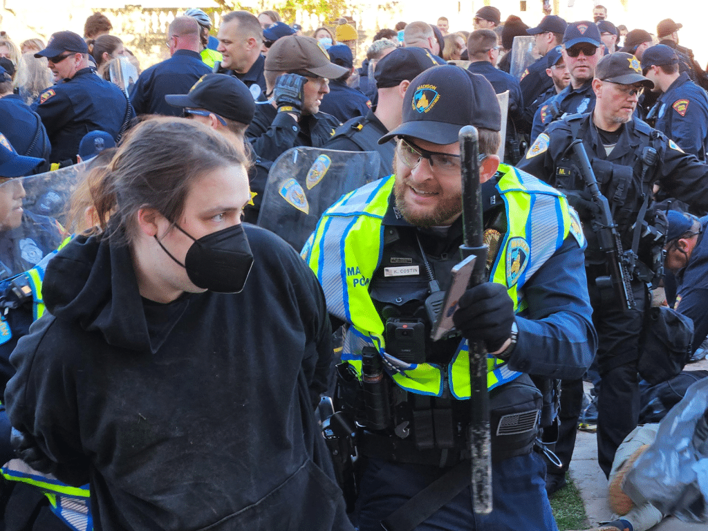 Photo from the police crackdown of the student protest encampment at UW-Madison on May 1, 2024. A UW-Madison police officer wearing a neon vest holds a baton and shows a smart-phone screen to a masked protestor in a black hoodie who is presumably handcuffed. The background shows a sea of officers and a general state of commotion.