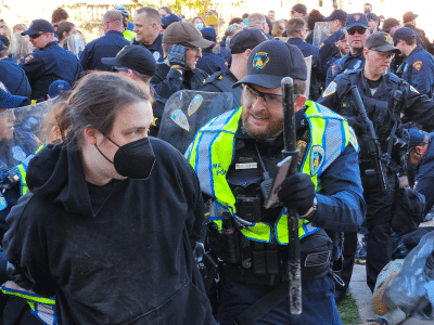 Photo from the police crackdown of the student protest encampment at UW-Madison on May 1, 2024. A UW-Madison police officer wearing a neon vest holds a baton and shows a smart-phone screen to a masked protestor in a black hoodie who is presumably handcuffed. The background shows a sea of officers and a general state of commotion.