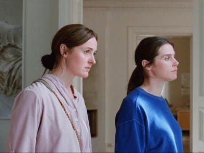 In a spacious and opulent house, two sisters stand at a medium shot in a sunlit room and look out to the right (through an unseen window). Both women have dark brown hair that is pulled back. They also both wear comfortable, long-sleeve clothing.