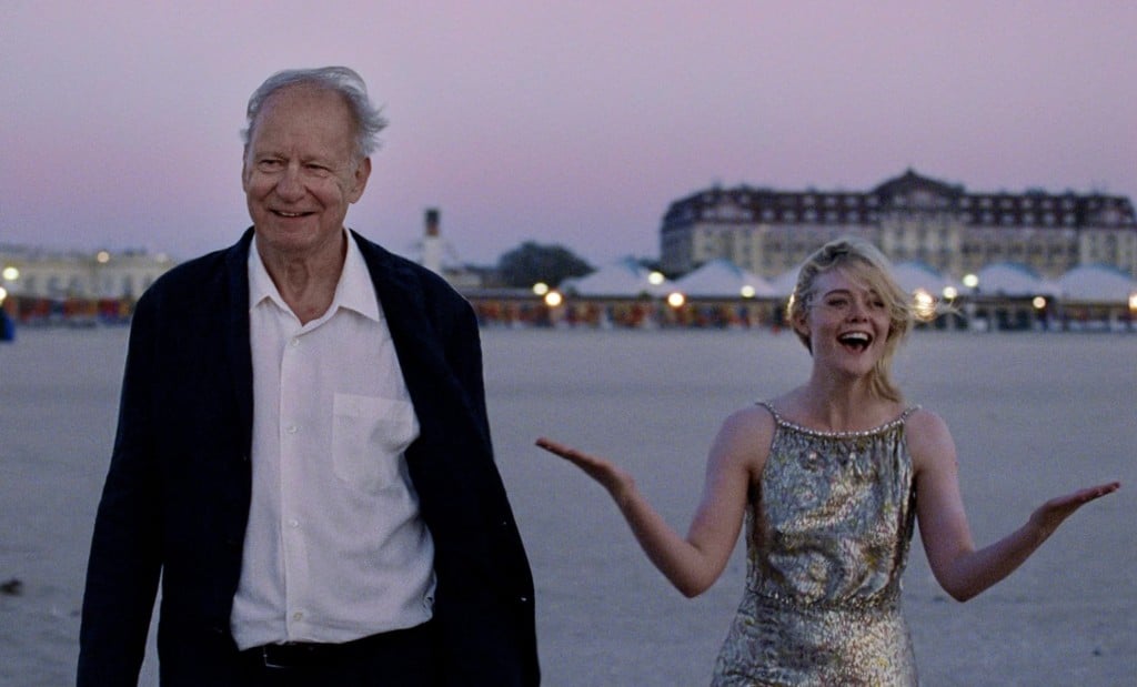 A promotional image from the film "Sentimental Value" shows an older man and younger woman standing on a beach at sunrise. The beach sands extend out behind them onto a plaza and hotel with some fluorescent lights still illuminated in the background. The figures, dressed rather formally in a suit and dress, look joyous, smiling widely in the foreground. The woman throws her arms out to the sides as if pleasantly surprised by something she sees off-screen.