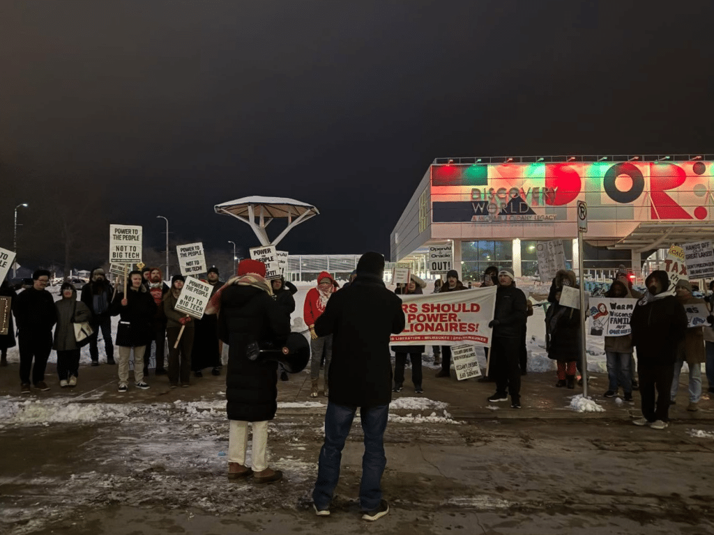 A protest during a winter night. The photo shows a couple dozen protestors bundled in heavy coats. Many hold white signs that read "Power To The People Not To Big Tech" in black lettering. A few others on the right address the resistance against AI and support of Wisconsin families. The boxy Discovery World Science and Technology Museum appears behind them to the right, illuminated in red and green above its sign.