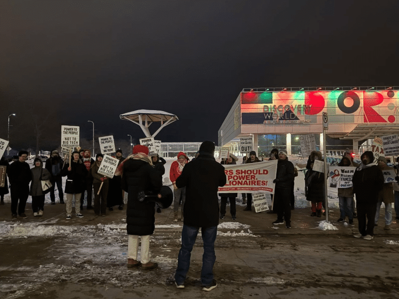 A protest during a winter night. The photo shows a couple dozen protestors bundled in heavy coats. Many hold white signs that read "Power To The People Not To Big Tech" in black lettering. A few others on the right address the resistance against AI and support of Wisconsin families. The boxy Discovery World Science and Technology Museum appears behind them to the right, illuminated in red and green above its sign.