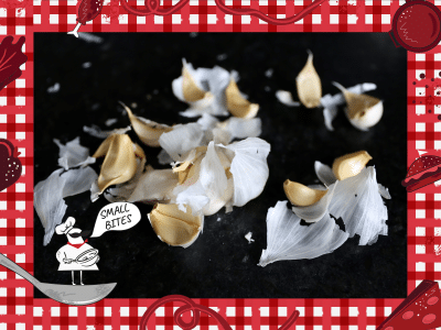 A close-up photo of several cloves of garlic, haphazardly peeled and scattered on a dark surface. Bordering this photo is a red and white checkered tablecloth illustration. In the lower left corner of this illustrated frame is a small chef with an oversized mustache standing on a spoon. The chef's speech bubble reads "Small Bites."