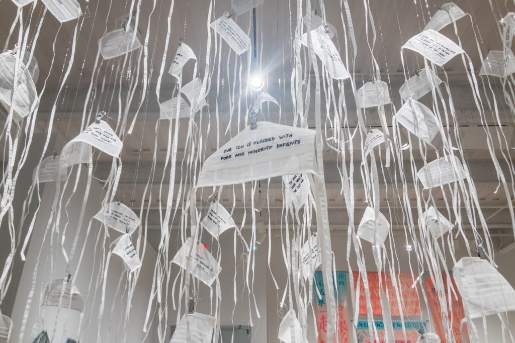 White organdy facemasks are suspended from the ceiling of an art gallery with word in black thread embroidered on them.