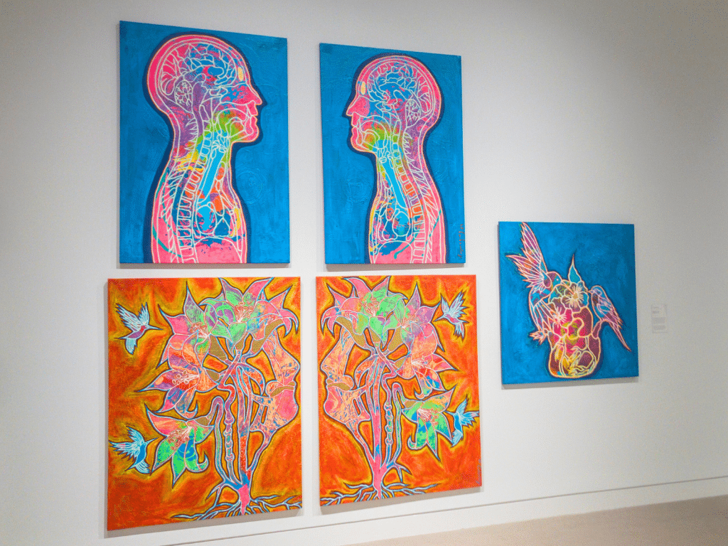 An exhibition of five large, brightly-colored paintings hanging on a white wall. The two uppermost paintings feature sky-blue backgrounds with anatomical renderings of human figures facing one another in profile. The bottom two paintings feature more abstract renderings of human heads facing one another. Blue hummingbirds are painted among the bright-orange background. The rightmost painting features a sky-blue background more birds withdrawing what appears to be nectar from a human heart.
