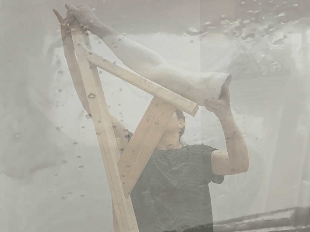 A still taken from Nicholas R. Wootton's experimental short "Liking This Angle" shows a woman holding up a plaster mold of a human arm on an angled wooden structure in an art studio. A second image of water streaking down a windshield is superimposed over it.