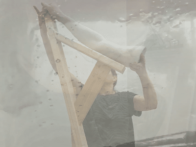 A still taken from Nicholas R. Wootton's experimental short "Liking This Angle" shows a woman holding up a plaster mold of a human arm on an angled wooden structure in an art studio. A second image of water streaking down a windshield is superimposed over it.