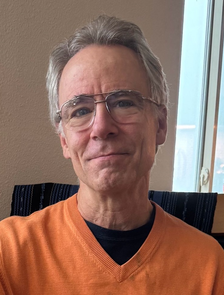 A headshot of the filmmaker Nicholas R. Wootton. He smiles slightly facing the camera, and wears glasses and an orange v-neck shirt.