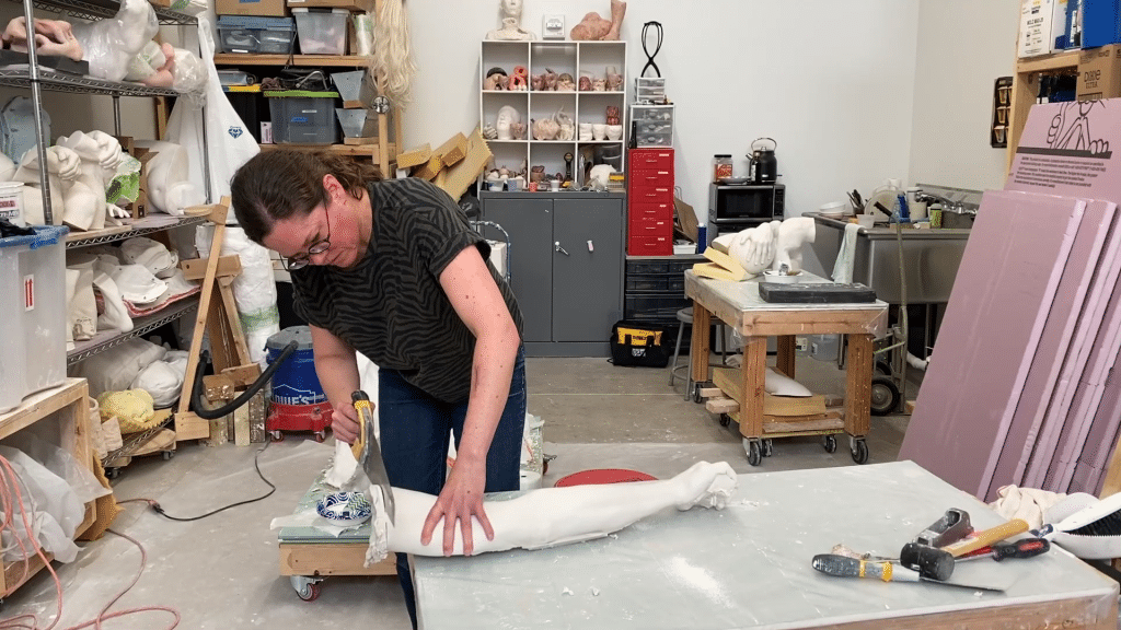 A still from the experimental short "Liking This Angle" shows a spacious art studio with various sculptures in the background on shelving units. In the foreground, the artist Christina A. West holds a metal saw in her right hand, while holding a plaster mold of a human arm in her left hand that's pressing it against the table for stability.