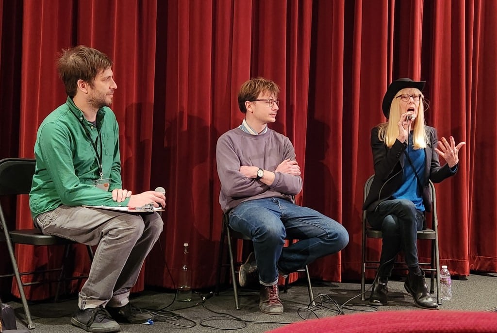 Three people sit on folding chairs at the front of a small auditorium stage with a red curtain. The woman on the right, wearing a black cowboy hat, holds a microphone and speaks as she gestures with her left hand. The two men to her left look towards her and listen attentively.