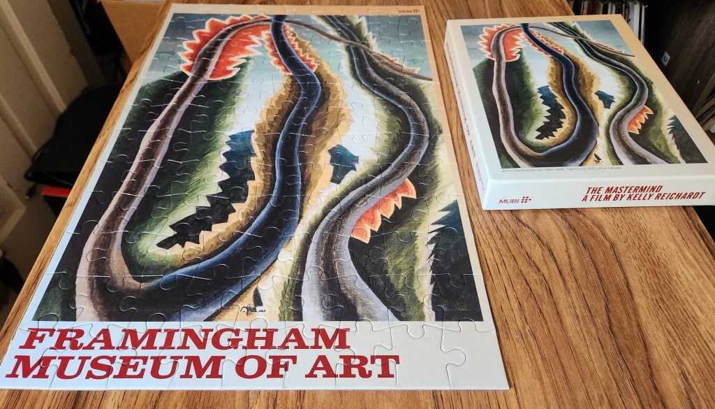 A jigsaw puzzle of a painting is completed and displayed on the surface of a wooden table. The painting—"Tree Forms" by Arthur Dove—shows a labyrinth of overlapping branches and foliage that are rendered in long, slender columns and jagged edges.