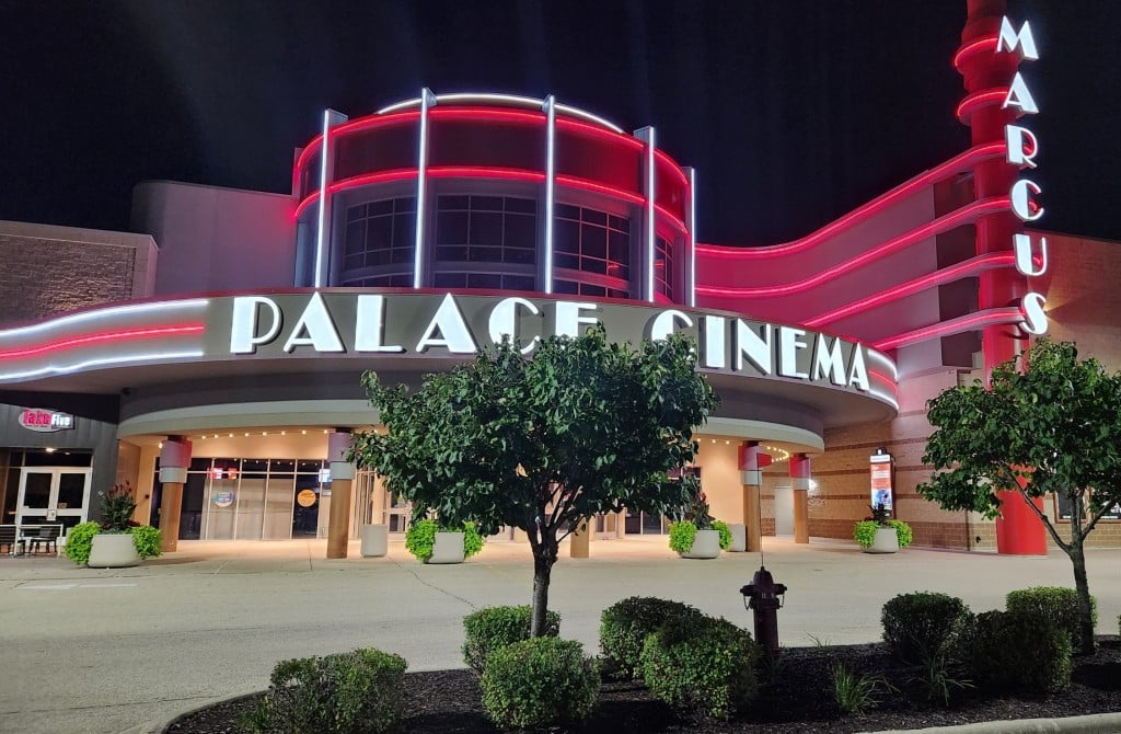 Photo taken from the parking lot of a neon-lit movie theater entrance that says "PALACE CINEMA" in white capital letters below red horizontal lines. To the right, a "MARCUS" sign in similar letting is positioned vertically, perpendicular to the main entrance.