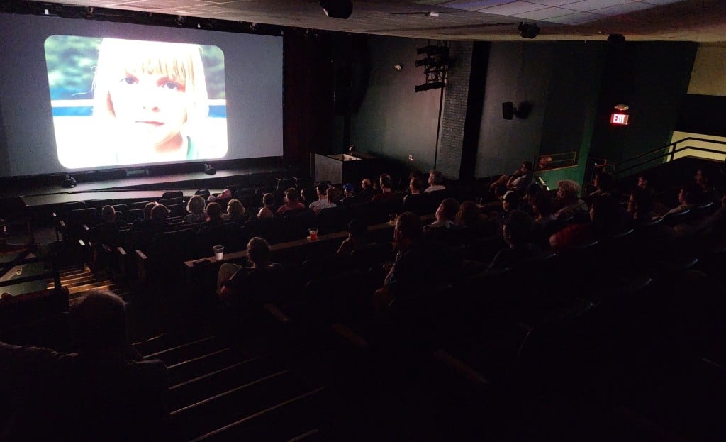 Photo of a darkened movie theater at an angle captures a crowd of people watching the illuminated screen at the top left, which shows a close-up of the face of Arabella Olivia Clark as Addie in the short film "Deuce."