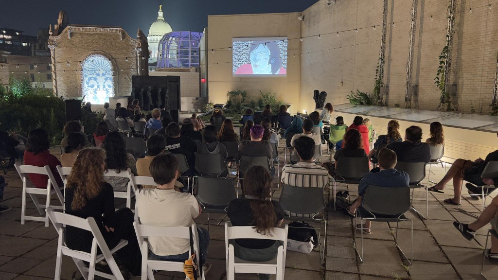 An outdoor sculpture garden space is populated with a few dozen people on various chairs. They sit in the partly illuminated space at night and stare ahead at a projection screen on the far wall.