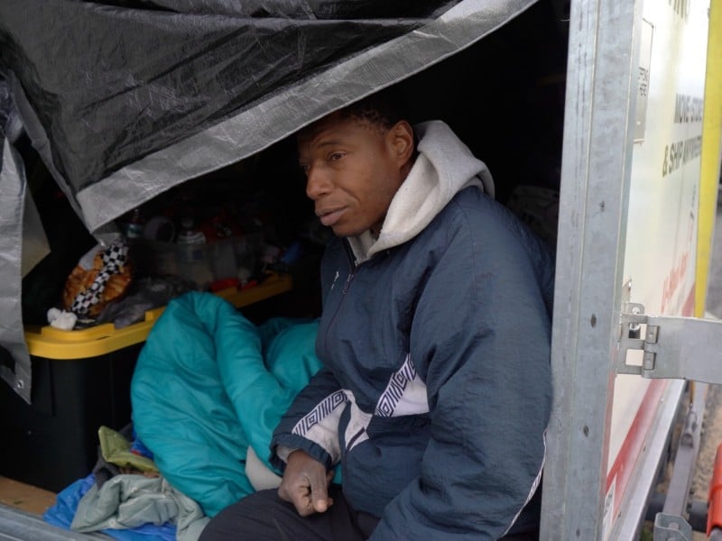 Photo of a Black man, Reggie, wearing a navy-blue windbreaker over a grey sweatshirt hoodie. He sits on the edge of a moving truck's deck that is partly covered by a black tarp. Inside the truck are several miscellaneous items like a teal blanket and black and yellow storage container.