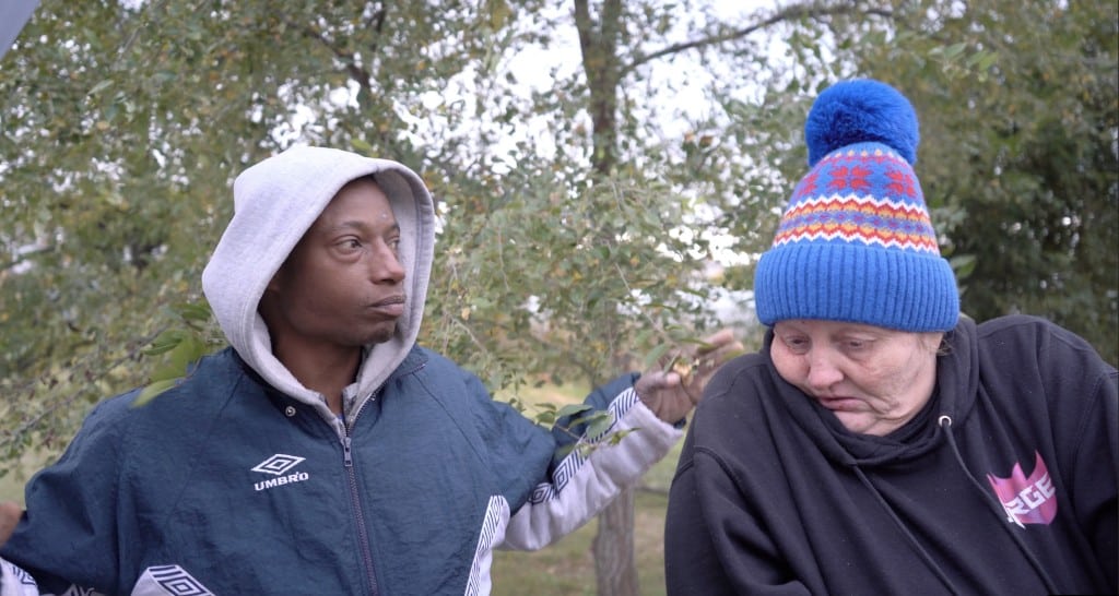 Candid photo of a Black man and a white woman standing outside in front of a tree. The man (on the left) wears a navy-blue windbreaker over a grey sweatshirt hoodie. The woman (on the right) wears a black sweatshirt hoodie and a blue winter hat with a colorful pattern design. Their heads are both turned away from the camera.