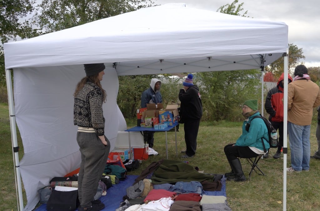 Photo of a white tent pitched outside on an overcast day that contains food and clothing items. Clothing sits on blankets in the grass and food on a blue folding table. Several people sit and stand around in warm-weather clothing, including the two subjects of this article- Reggie and Anne- centered in the middleground.