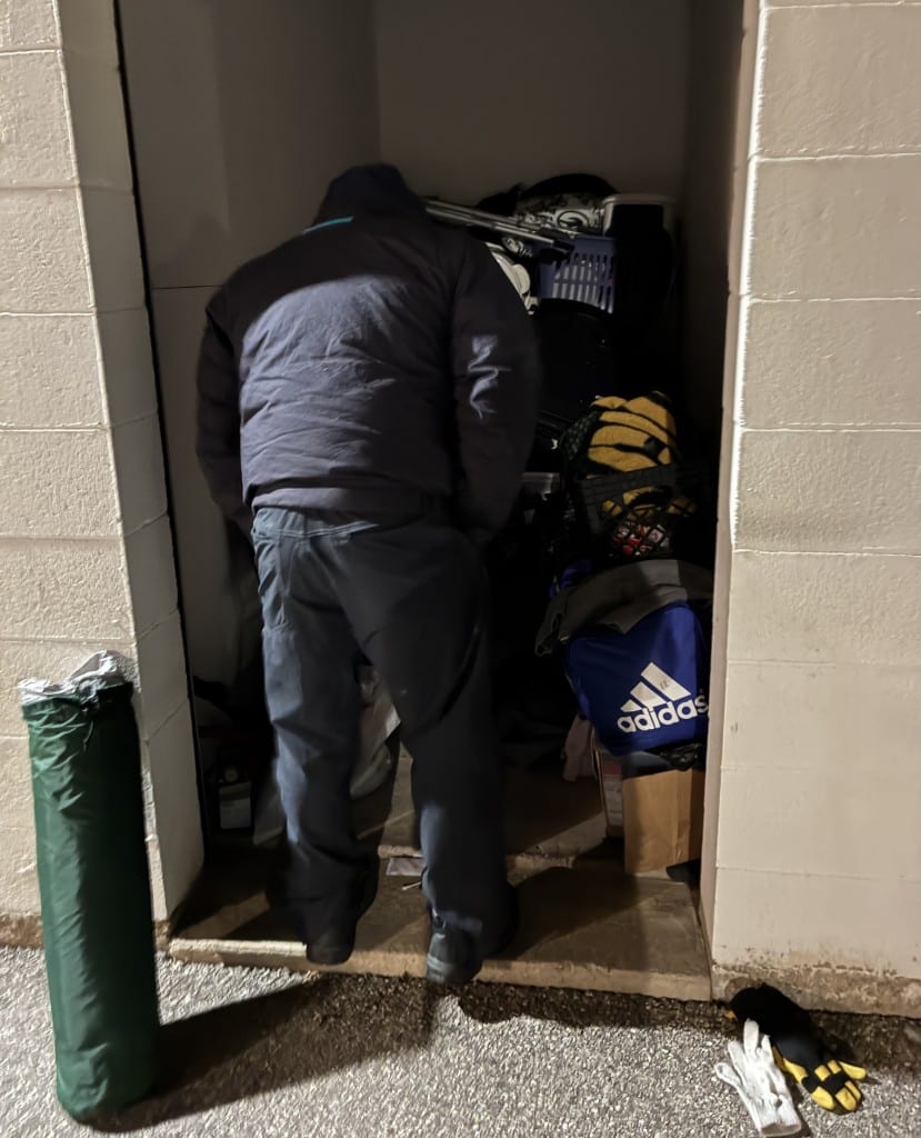 Photo of a man with his back to the camera looking in a tan-colored storage unit. He wears a black coat and pants. A green lawn chair is rolled up in a bar behind him against the wall.