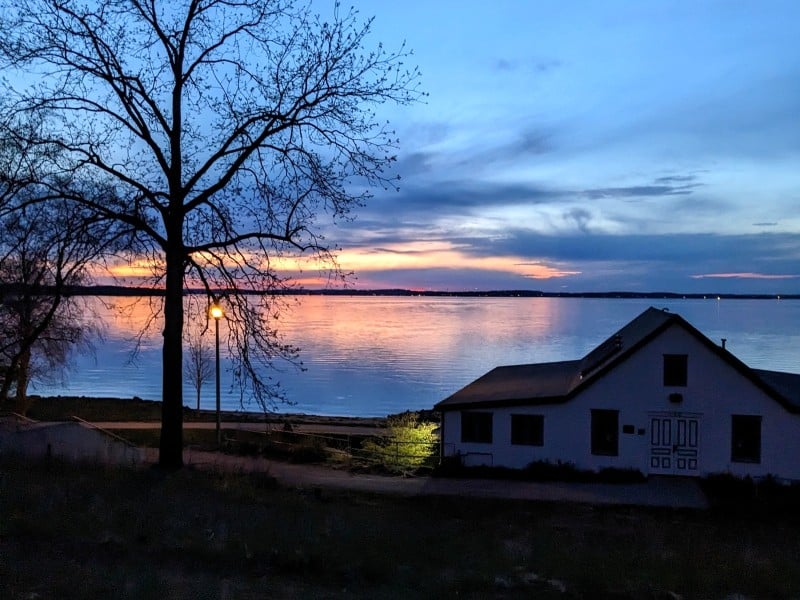A long shot photo of Lake Mendota at either dawn or dusk time, in limited light. The sky has a pretty mix of dark and light blue colors amid the yellow-orange of the sun near the horizon line. A large budding tree stands in the middleground next to an illuminated lamppost.