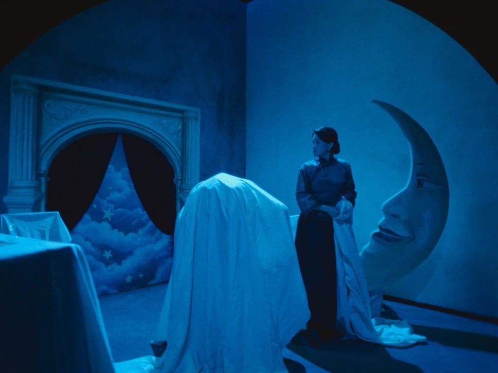 A still frame from the film "Resurrection" (2025) shows a corner stage area bathed in a French navy blue-grey light. The stage is littered with oversized props (some covered by white sheets) that recall the films of Georges Méliès from the late 19th and early 20th century—including a crescent moon with a face and a fireplace with a black curtain that is partly pulled back to reveal a starry and cloudy sky. A female actor wears a traditional Chinese dress and looks off to the left in the foreground.