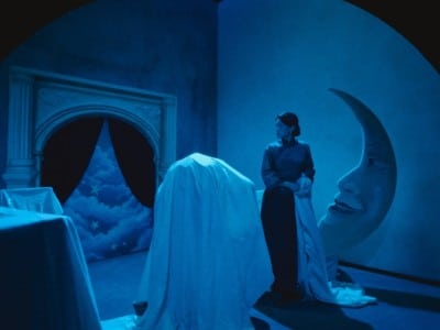 A still frame from the film "Resurrection" (2025) shows a corner stage area bathed in a French navy blue-grey light. The stage is littered with oversized props (some covered by white sheets) that recall the films of Georges Méliès from the late 19th and early 20th century—including a crescent moon with a face and a fireplace with a black curtain that is partly pulled back to reveal a starry and cloudy sky. A female actor wears a traditional Chinese dress and looks off to the left in the foreground.