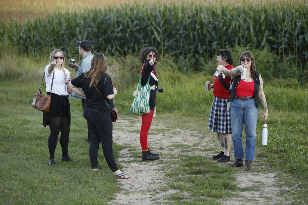 The members of Crush Scene and two-thirds of Holly And The Nice Lions all stand on a vineyard path at The Vines in Sauk City, WI. From left to right: Holly Trasti, Travis Pashek, Rachael Guertin, Janet Kolterman, Tasha Spencer, and Madalyn Rowell. Guertin and Pashek have their backs turned to the camera, admiring the sweeping view. The remaining subjects are all pointing directly at the camera and smiling.