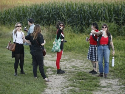 The members of Crush Scene and two-thirds of Holly And The Nice Lions all stand on a vineyard path at The Vines in Sauk City, WI. From left to right: Holly Trasti, Travis Pashek, Rachael Guertin, Janet Kolterman, Tasha Spencer, and Madalyn Rowell. Guertin and Pashek have their backs turned to the camera, admiring the sweeping view. The remaining subjects are all pointing directly at the camera and smiling.