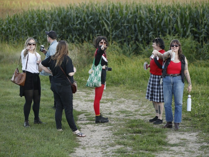 The members of Crush Scene and two-thirds of Holly And The Nice Lions all stand on a vineyard path at The Vines in Sauk City, WI. From left to right: Holly Trasti, Travis Pashek, Rachael Guertin, Janet Kolterman, Tasha Spencer, and Madalyn Rowell. Guertin and Pashek have their backs turned to the camera, admiring the sweeping view. The remaining subjects are all pointing directly at the camera and smiling.