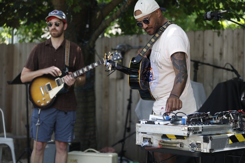 Def Sonic perform as part of Make Music Madison. They're performing outside on a warm summer day. One member is out of focus to the far left in the background, the other is in focus, holding an acoustic guitar and looking down while he manipulates an effects board.