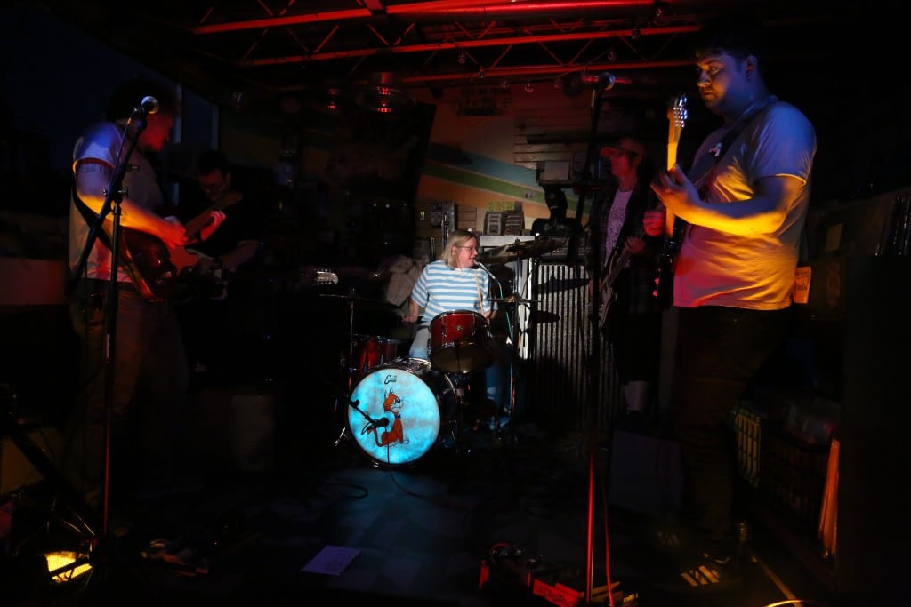 Heather The Jerk performs in a record store and novelty retro toy shop. Drummer Heather Sawyer is clearly lit and centered in the background, while her remaining four bandmates are shrouded in dark, shadowy light. They are split evenly to the left and the right.